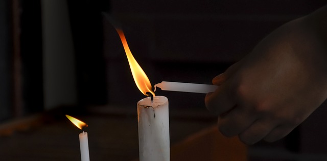 Bild von Pexels auf Pixabay 2 Kerzen brennen, eine Hand mit einer weiteren Kerze zündet die Kerze an einer der anderen beiden an. Der Hintergrund ist schwarz.